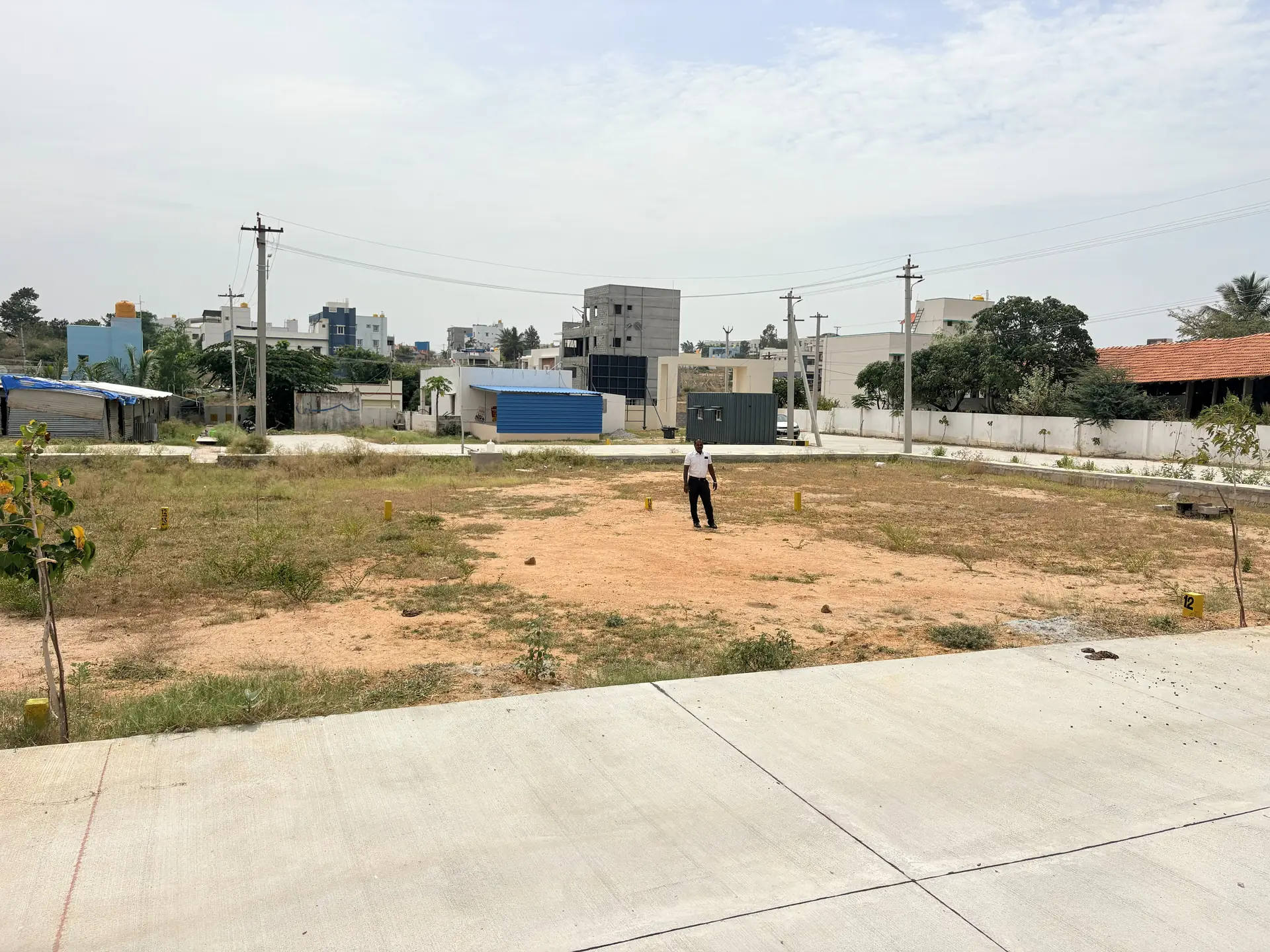A marked plot at Poes Garden beside a finished concrete road, with completed houses in the neighbourhood behind.