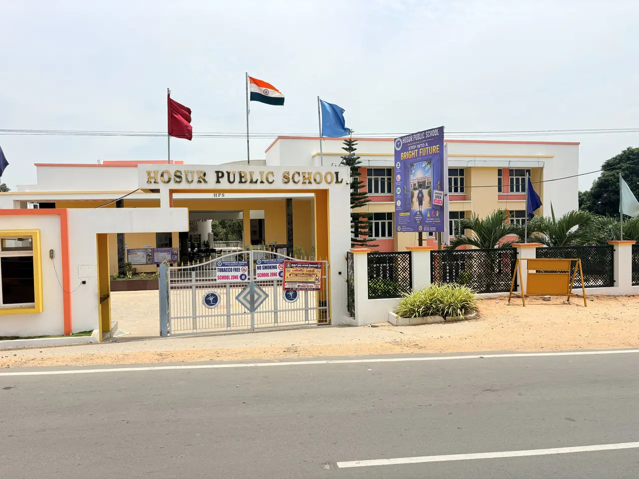 Hosur Public School seen from the main road, its name across the porch and flags flying above it.