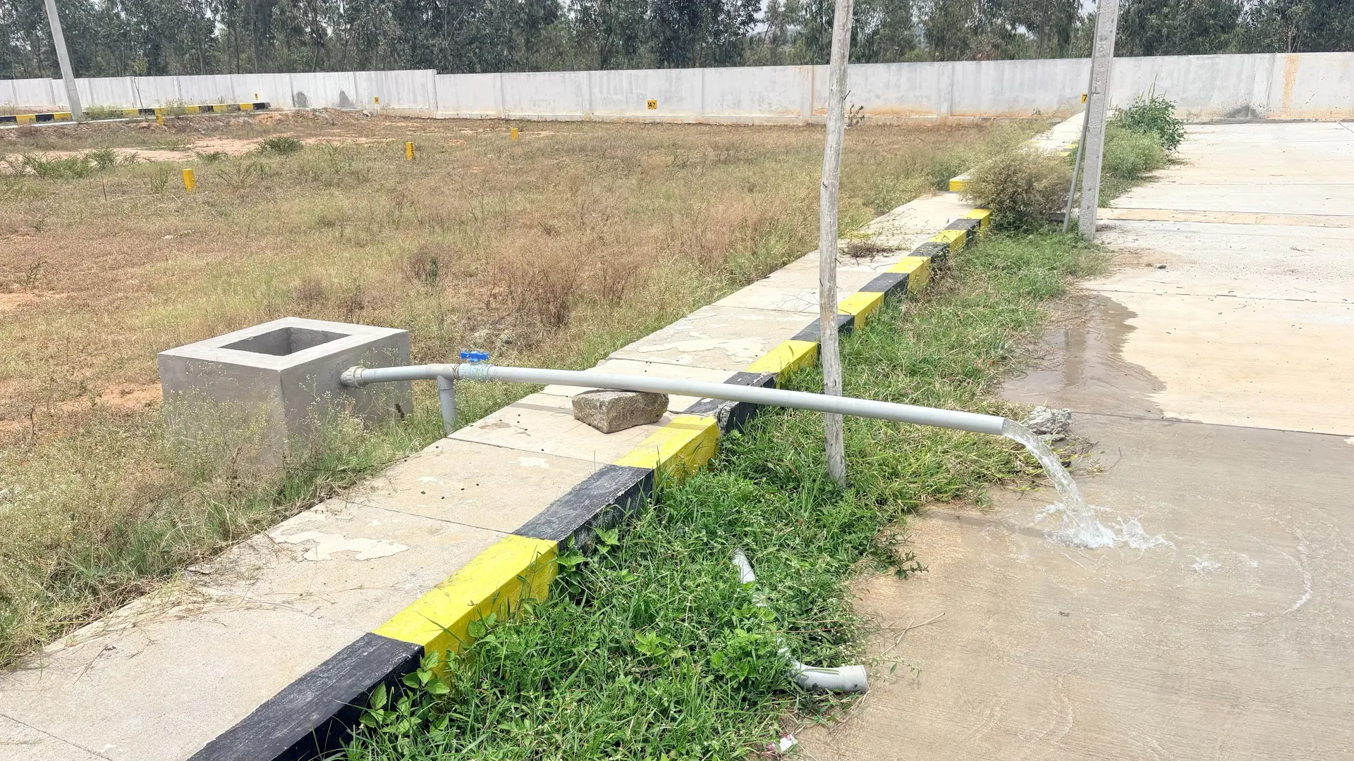Water running from a supply pipe beside a chamber at the edge of a plot, with the compound wall behind.